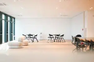 Spacious, brightly lit room with white walls, several black chairs around tables, and a white cushioned seat near large windows on the left.