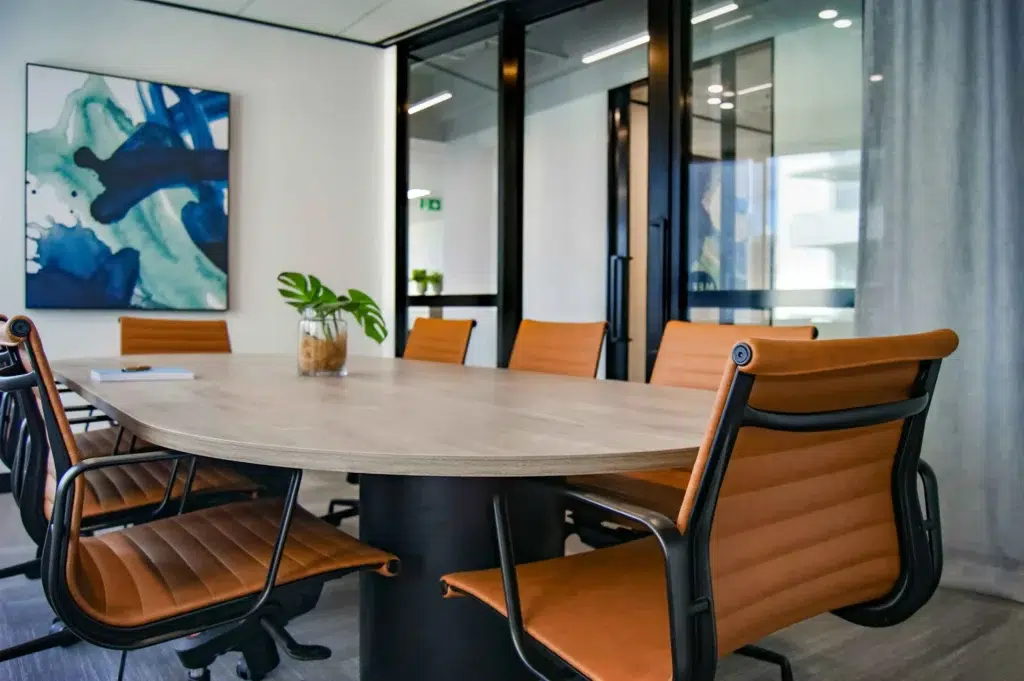 Modern conference room with a wooden oval table, surrounded by six brown leather chairs. Abstract blue artwork on the wall; vibrant and professional ambiance.